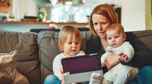 A mother watches a video on her iPad while holding her infant and toddler sitting on the couch.