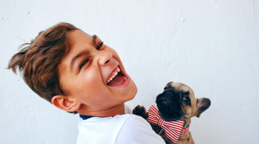 A little boy holds his dog, wearing a bow tie, and laughs.