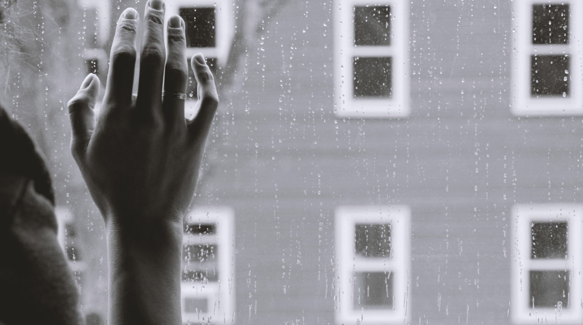 A person in greyscale stands at a window while it rains.