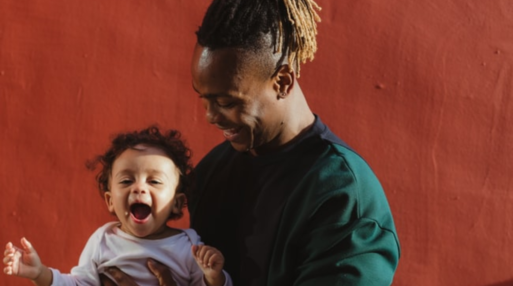 A father holds his child outside in a playful way while they both laugh against a red concrete wall.